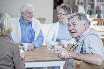 Amis parlant et souriant à table avec des boissons dans la maison de retraite . — Photo de stock