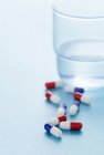 Червоні та сині капсули ліків розкидані на синьому фоні зі склянкою води поруч . — стокове фото