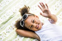 Vue aérienne de la petite fille d'âge préscolaire couchée sur une couverture dans le parc, souriant et agitant, portrait . — Photo de stock