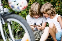 Niños jugando juego en el teléfono inteligente detrás de la bicicleta en el parque
. - foto de stock