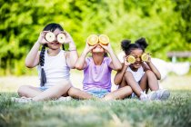 Elementary age girls sitting side by side in park and covering eyes with fruit slices. — Stock Photo
