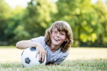 Boy holding soccer ball while lying on green grass in park and smiling, portrait. — Stock Photo