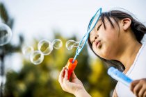 Low angle view of Asian girl blowing bubbles with bubble wand in park. — Stock Photo