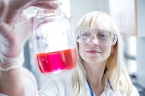 Cientista feminina vestindo óculos de proteção e segurando frasco com líquido rosa . — Fotografia de Stock