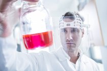 Científico masculino usando máscara protectora y sosteniendo frasco con líquido rosa . - foto de stock