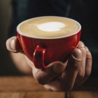 Professional barista holding in hand red coffee cup with heart latte art surface. — Stock Photo