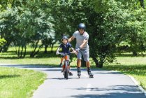 Active senior on roller skates teaching grandson riding bicycle in park. — Stock Photo