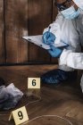 Forensics expert in protective suit writing in clipboard collecting evidence from crime scene. — Stock Photo