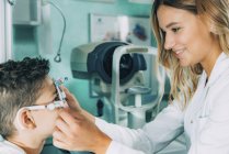 Oftalmólogo ayudando a niño a usar anteojos mientras examinan los ojos en la clínica . - foto de stock