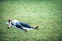 Mujer haciendo yoga y meditando en posición de cadáver shavasana en estera en parque . - foto de stock