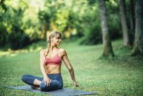 Young woman doing yoga and twisting from lotus position on mat in park. — Stock Photo
