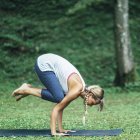 Mujer joven haciendo yoga, practicando la posición de cuervo bakasana en la estera en el parque . - foto de stock