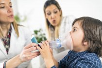 Pulmonologist helping little boy with inhaler, mother in background. — Stock Photo