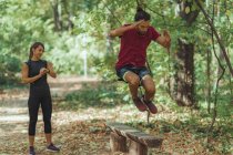 Young man jumping over wooden bench while training with personal trainer in park. — Stock Photo