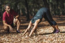 Woman in downward-facing dog yoga pose while training with personal trainer in park. — Stock Photo