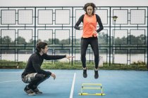 Sporty woman jumping over hurdle with fitness coach assistance. — Stock Photo