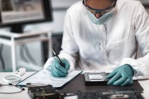 Female digital forensic expert examining computer hard drive and taking notes in police science laboratory. — Stock Photo