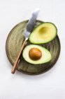 Avocado cut in half with knife on round plate on white background. — Stock Photo