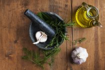 Mortar and pestle with garlic and herbs and olive oil on wooden table, top view. — Stock Photo