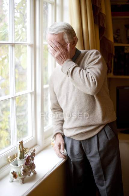 Старший чоловік з долонею на обличчі стоїть біля вікна в домашньому інтер'єрі . — стокове фото