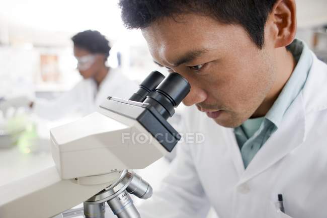 Male biologist working with microscope with colleague in background — Stock Photo
