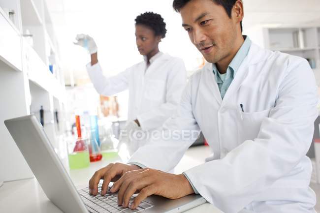 Chemiker arbeitet im Labor mit Laptop und Kollege im Hintergrund. — Stockfoto