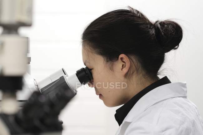 Técnico femenino en bata blanca usando microscopio . - foto de stock