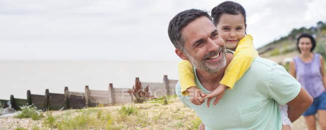 Father giving daughter piggyback on beach with mother. — Stock Photo