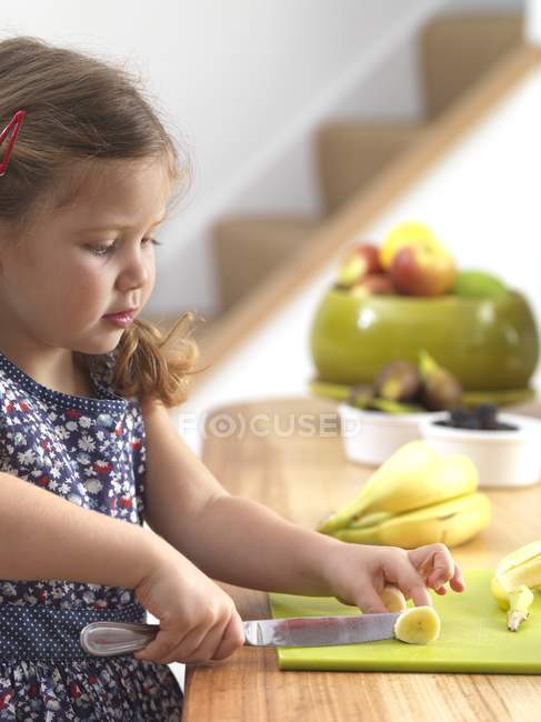 Niña preescolar cortando plátano en la cocina . - foto de stock