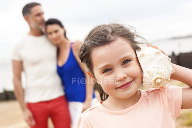 Young girl listening to seashell with parents in background. — Stock Photo