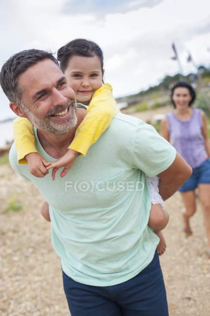 Father giving daughter piggyback on beach with mother in background. — Stock Photo