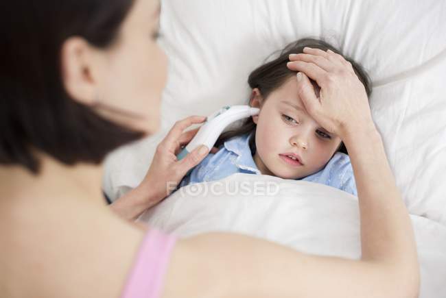 Madre tomando temperatura hija con la mano y el dispositivo en la cama . - foto de stock