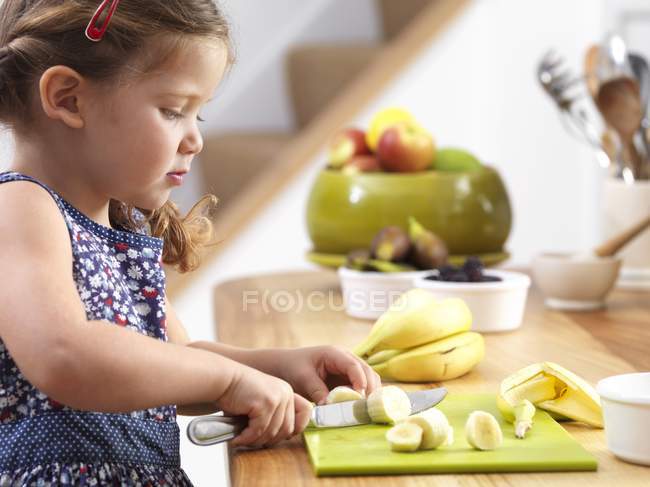 Préscolaire fille coupe banane dans la cuisine . — Photo de stock