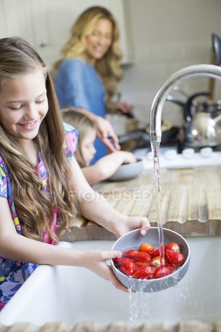 Chica lavando fresas frescas en fregadero de cocina con madre y hermana en el fondo . - foto de stock