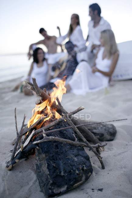 Group of young people relaxing at beach with bonfire in foreground. — Stock Photo