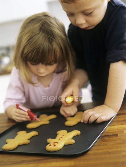 Frères et sœurs d'âge préscolaire décorant des biscuits fraîchement cuits au four avec du glaçage . — Photo de stock