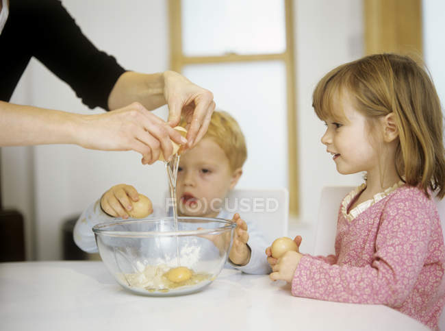 Los niños viendo como la madre rompe el huevo en un tazón . - foto de stock