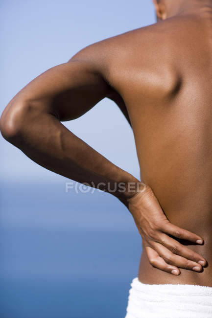 Man holding painful lower back on beach, rear view. — Stock Photo