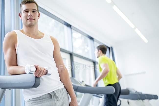 Mann hält Hantel in der Hand und lehnt sich an Laufband im Fitnessstudio. — Stockfoto