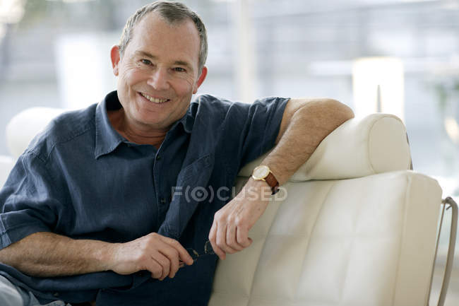 Hombre sosteniendo gafas y mirando en la cámara mientras se sienta y se apoya en el sofá . - foto de stock