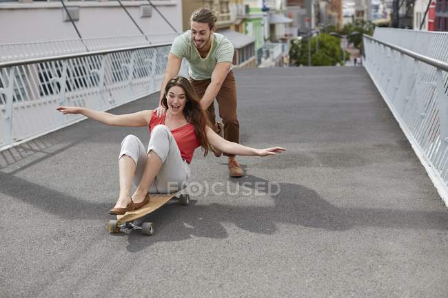 Mulher sentada no skate com homem empurrando na rua . — Fotografia de Stock