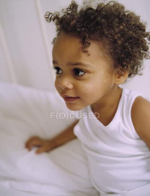 Niño pequeño con rizos vistiendo camiseta y sentado en la cama . - foto de stock