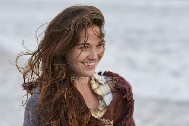 Mujer joven con el pelo desordenado de pie en la playa y sonriendo . - foto de stock