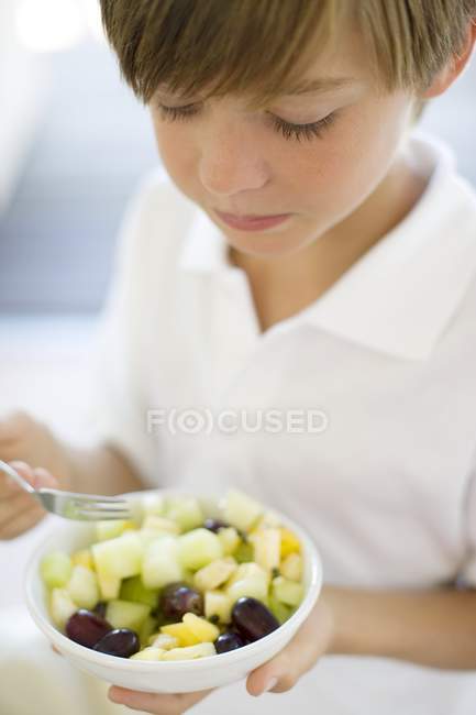 Menino da idade elementar comendo salada de frutas em tigela . — Fotografia de Stock