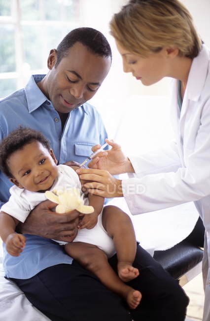Female doctor giving infant boy injection while baby sitting on father lap. — Stock Photo