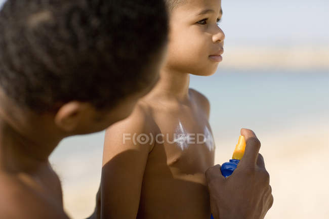 Man spraying sun cream onto son at beach. — Stock Photo