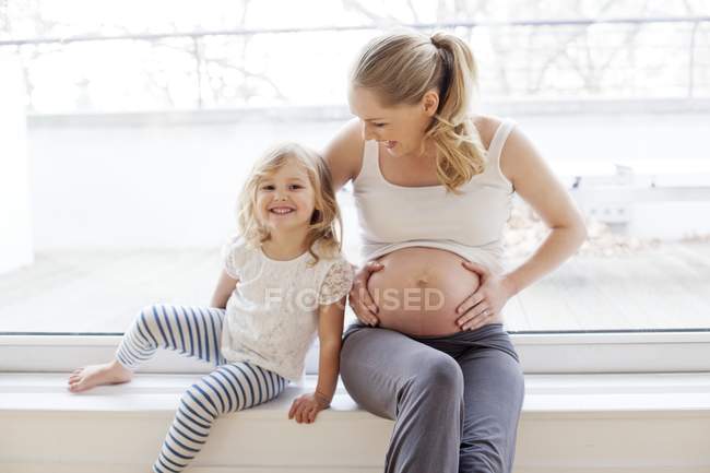 Pregnant mother touching tummy with daughter on window sill. — Stock Photo