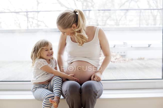 Kleines Mädchen berührt schwangeren Mutterbauch auf Fensterbank. — Stockfoto