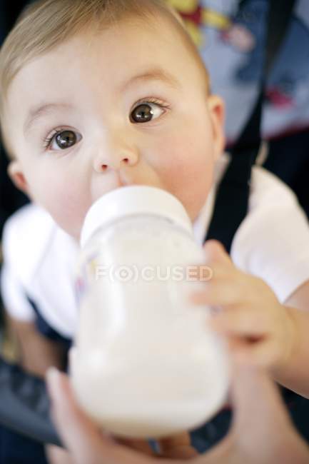 Female hand helping baby boy drinking bottle of milk in safety chair. — Stock Photo