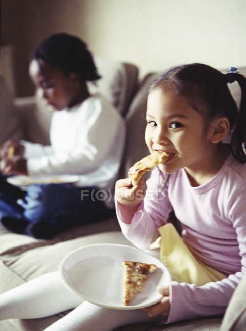 Elemental chica edad tomando mordida de rebanada de pizza con hermano en el fondo . - foto de stock